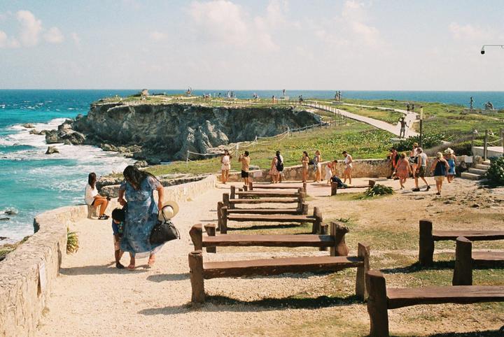 A group of people are walking along a path next to a rocky coastline with turquoise water.