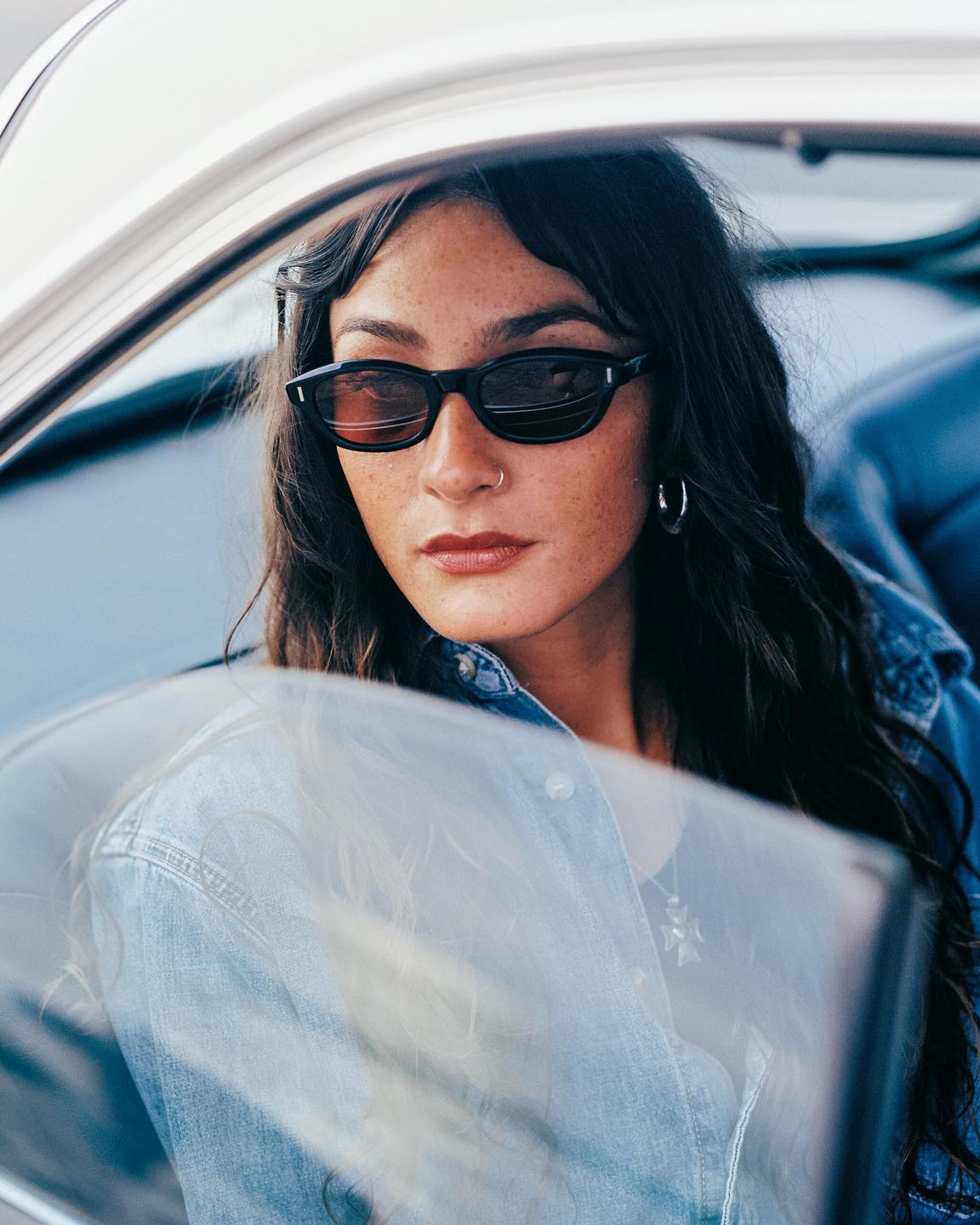 A woman with long hair and sunglasses looks out of a car window.