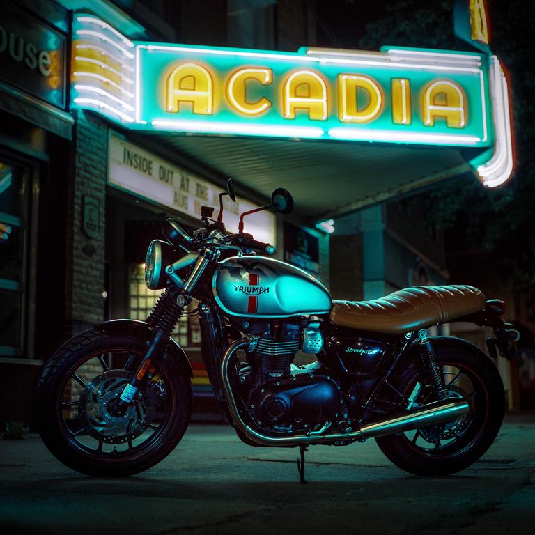 A motorcycle is parked on a street at night under a neon sign that reads "Acadia."