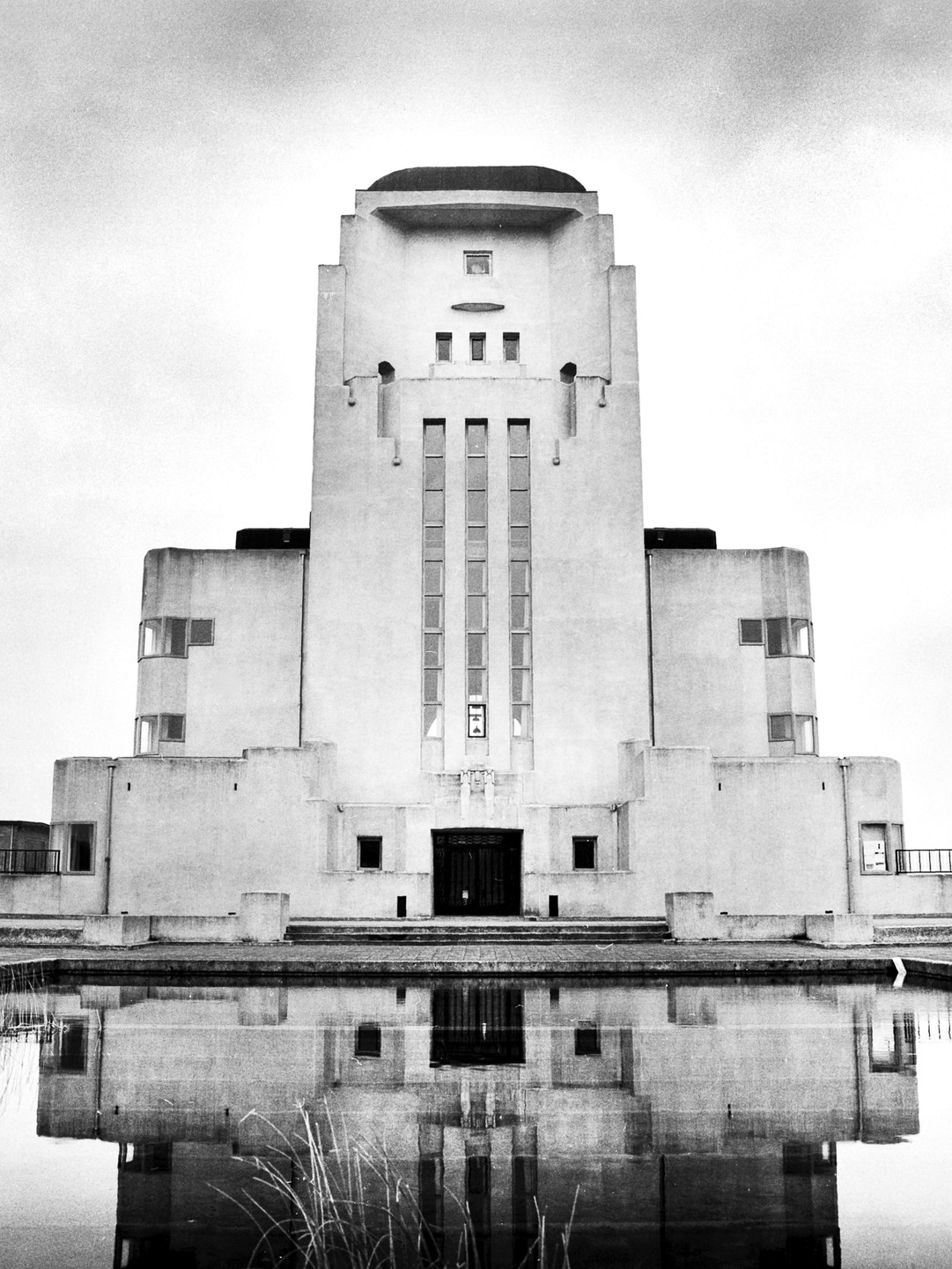 A tall, art deco building with a dark entrance is reflected in a body of water with some reeds.