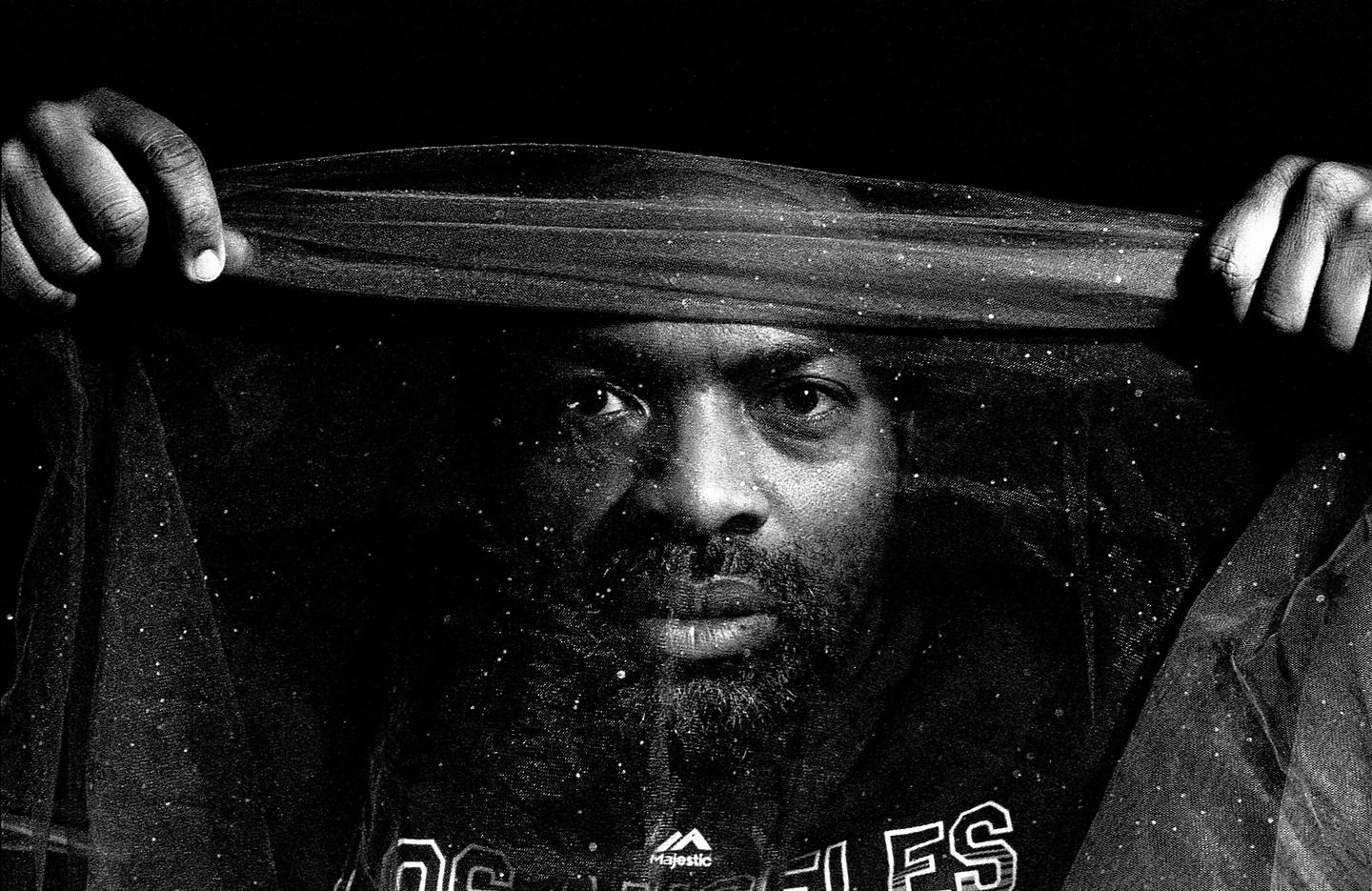 A man with a beard holds a sheer, glittery fabric over his head, looking directly at the viewer.