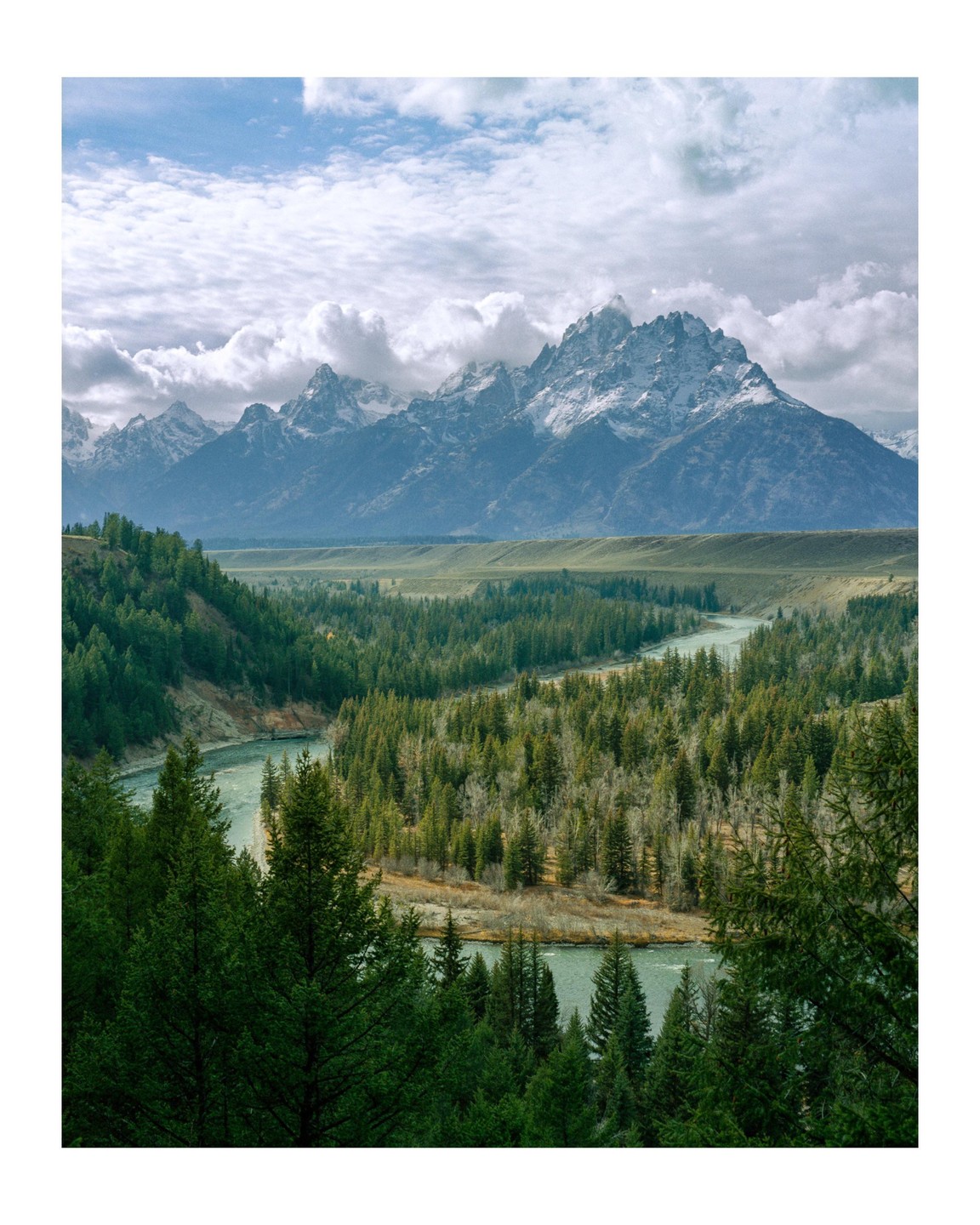 A wide river winds through a dense forest with snow-capped mountains under a cloudy sky in the background.