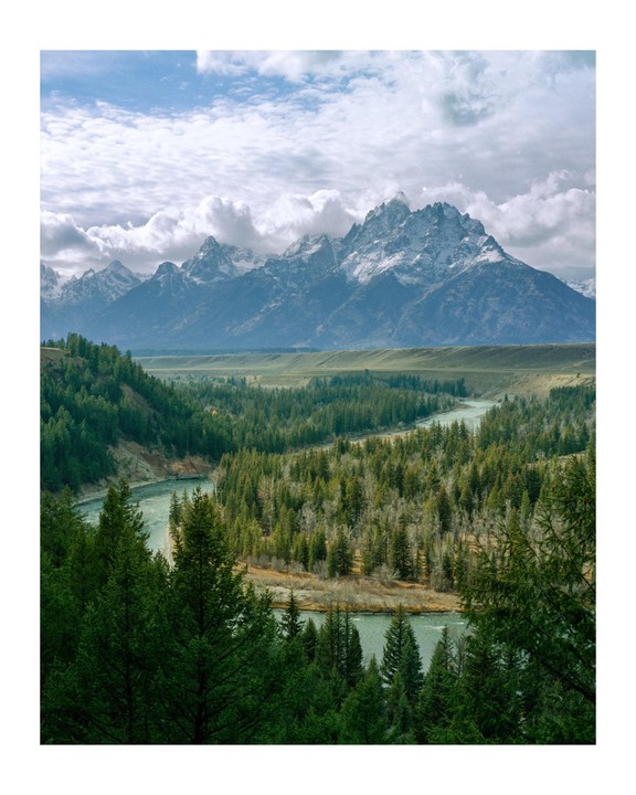 A wide river winds through a dense forest with snow-capped mountains under a cloudy sky in the background.