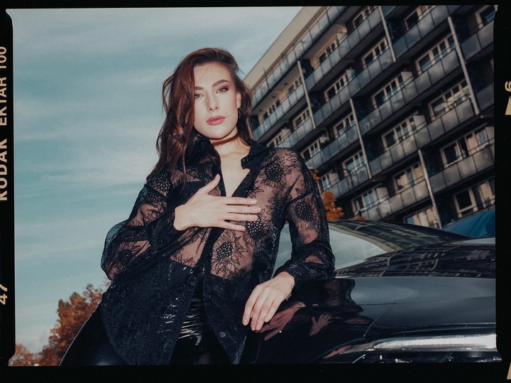 A woman in a sheer black lace shirt leans on a car with an apartment building in the background.
