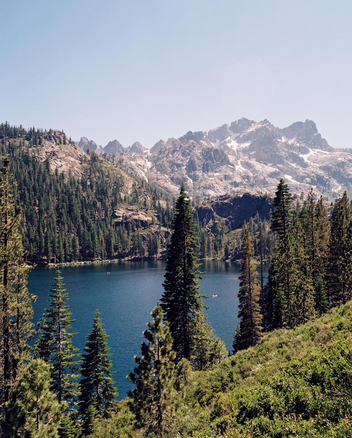 A deep blue lake is surrounded by a dense forest and snow-capped mountains under a clear sky.
