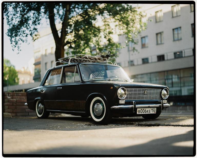 A black car with a roof rack is parked on a street next to a building and a tree.