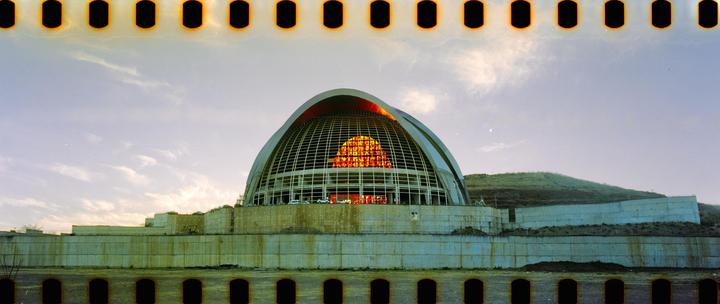 A large domed building with a grid-like facade and glowing orange windows sits on a concrete base next to a grassy hill under a cloudy sky.