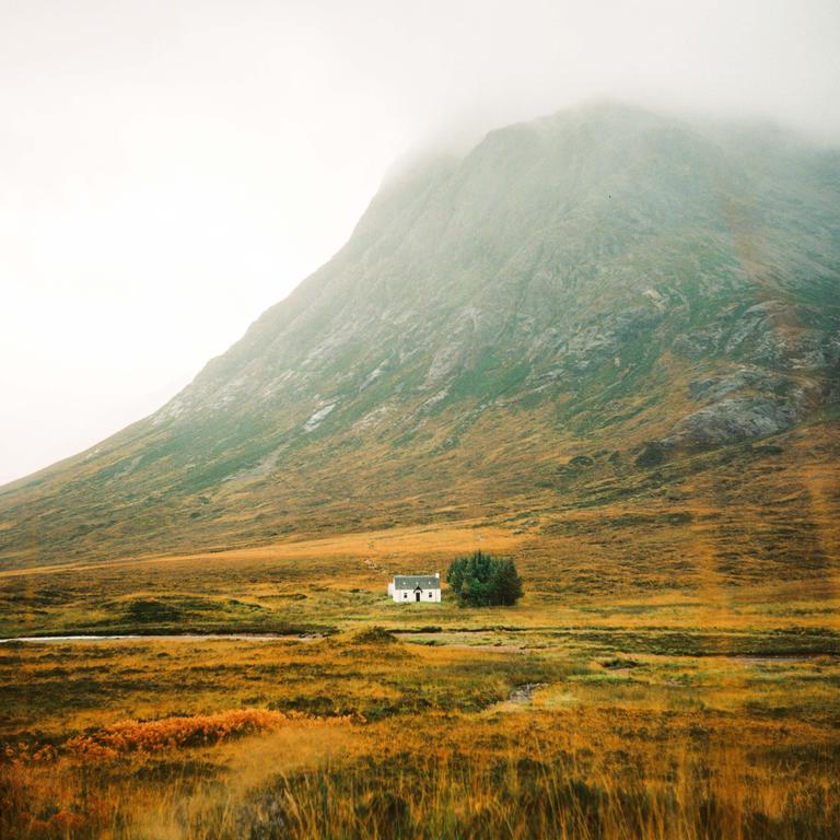A small white house sits in a field of golden grass at the base of a large mountain shrouded in fog.