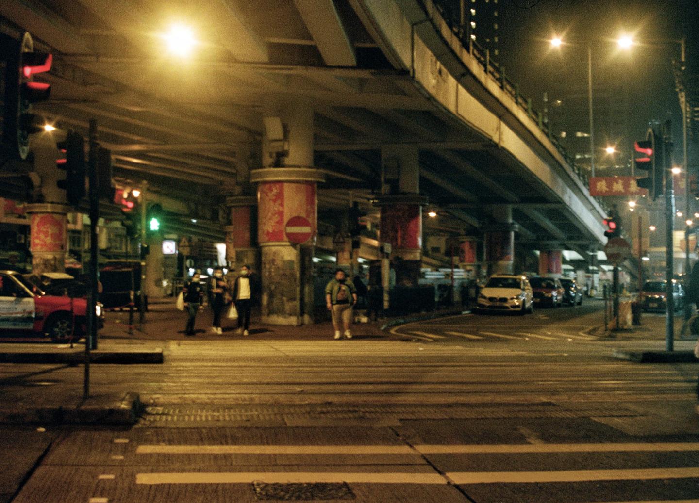 A street at night is illuminated by streetlights and traffic lights, with an overpass above and cars and people below.