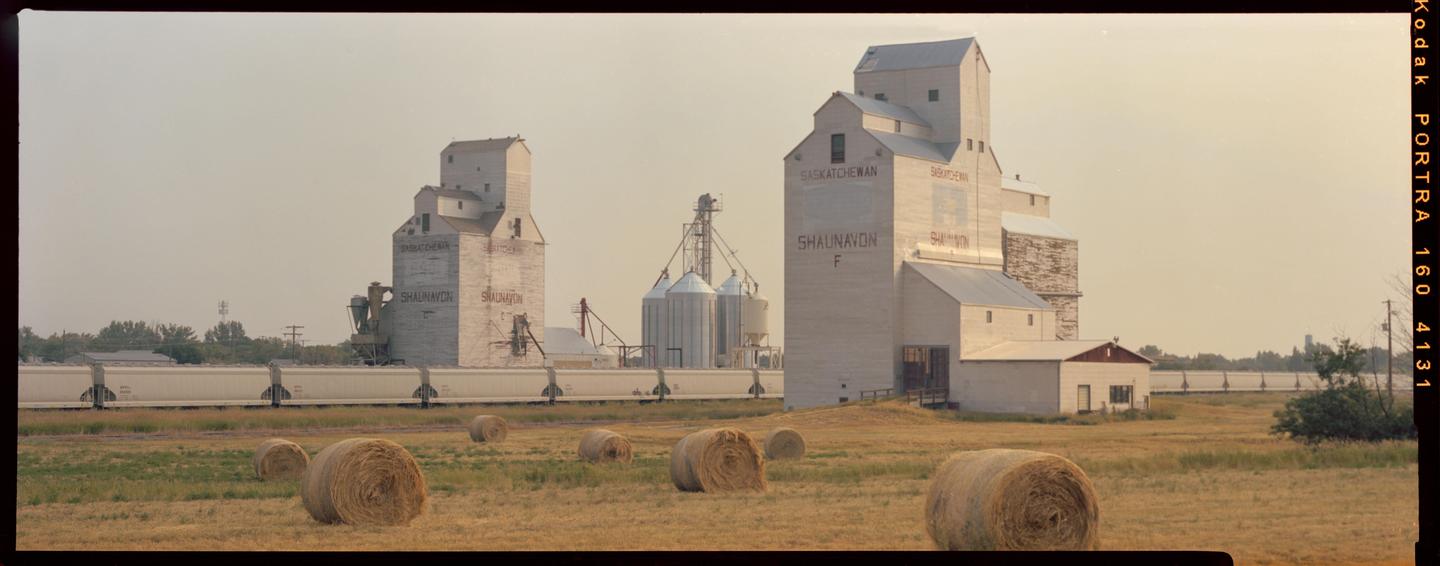 Two large grain elevators stand in a field with hay bales and a train passing by.