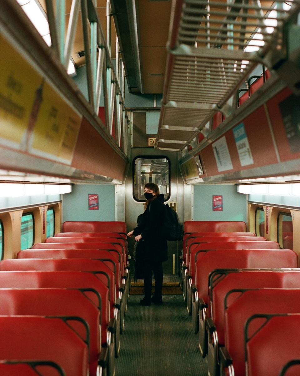 A woman wearing a face mask stands in the aisle of an empty train car with red seats.
