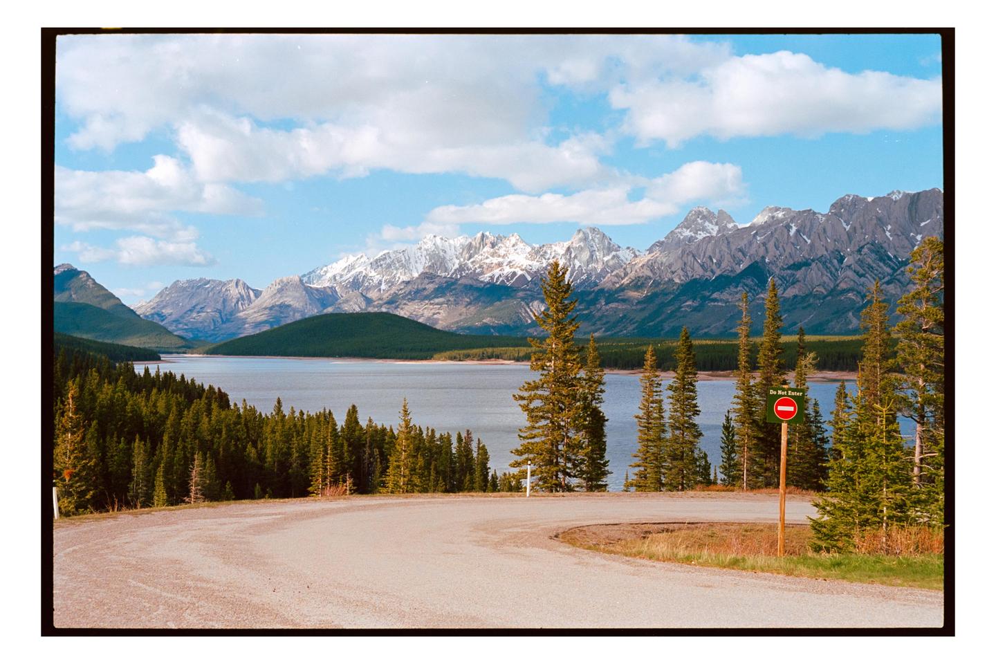 A winding road overlooks a lake surrounded by a forest with snow-capped mountains in the distance under a blue sky with clouds.
