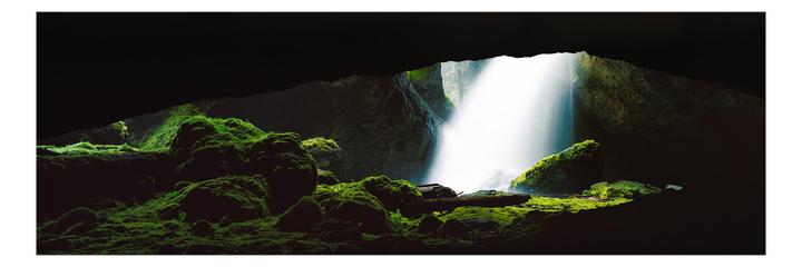 A bright waterfall cascades into a dark cave, illuminating moss-covered rocks and logs.