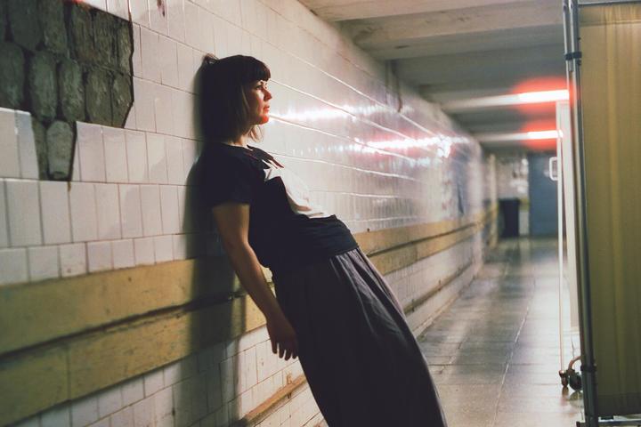 A woman leans against a tiled wall in a long, narrow hallway with red lights.