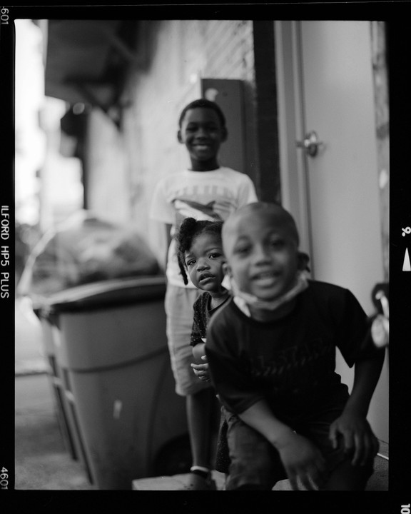Three children stand in a line on a street in Brooklyn.