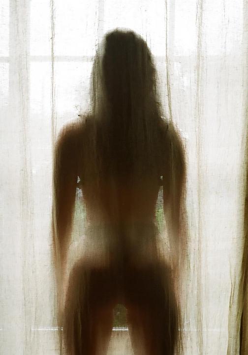 A woman stands behind a sheer curtain, her silhouette visible against the bright window.