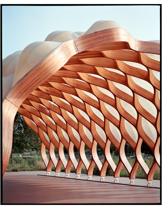 A wooden lattice structure with a wavy roof stands over a paved area, casting shadows on the ground.