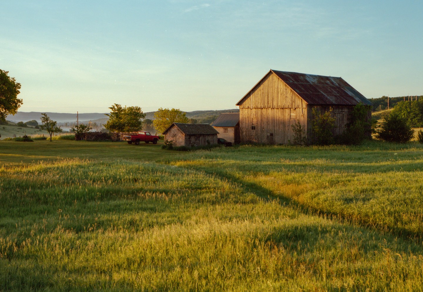 A red truck is parked next to a small shed and a large barn in a field at sunrise.