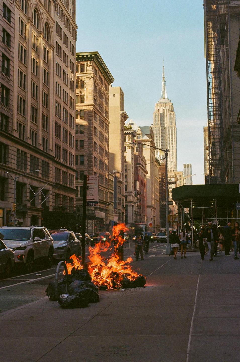 A pile of burning garbage bags sits on a new york city street with the empire state building in the background.