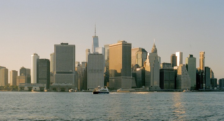 A ferry boat crosses a body of water in front of a city skyline with many tall buildings.