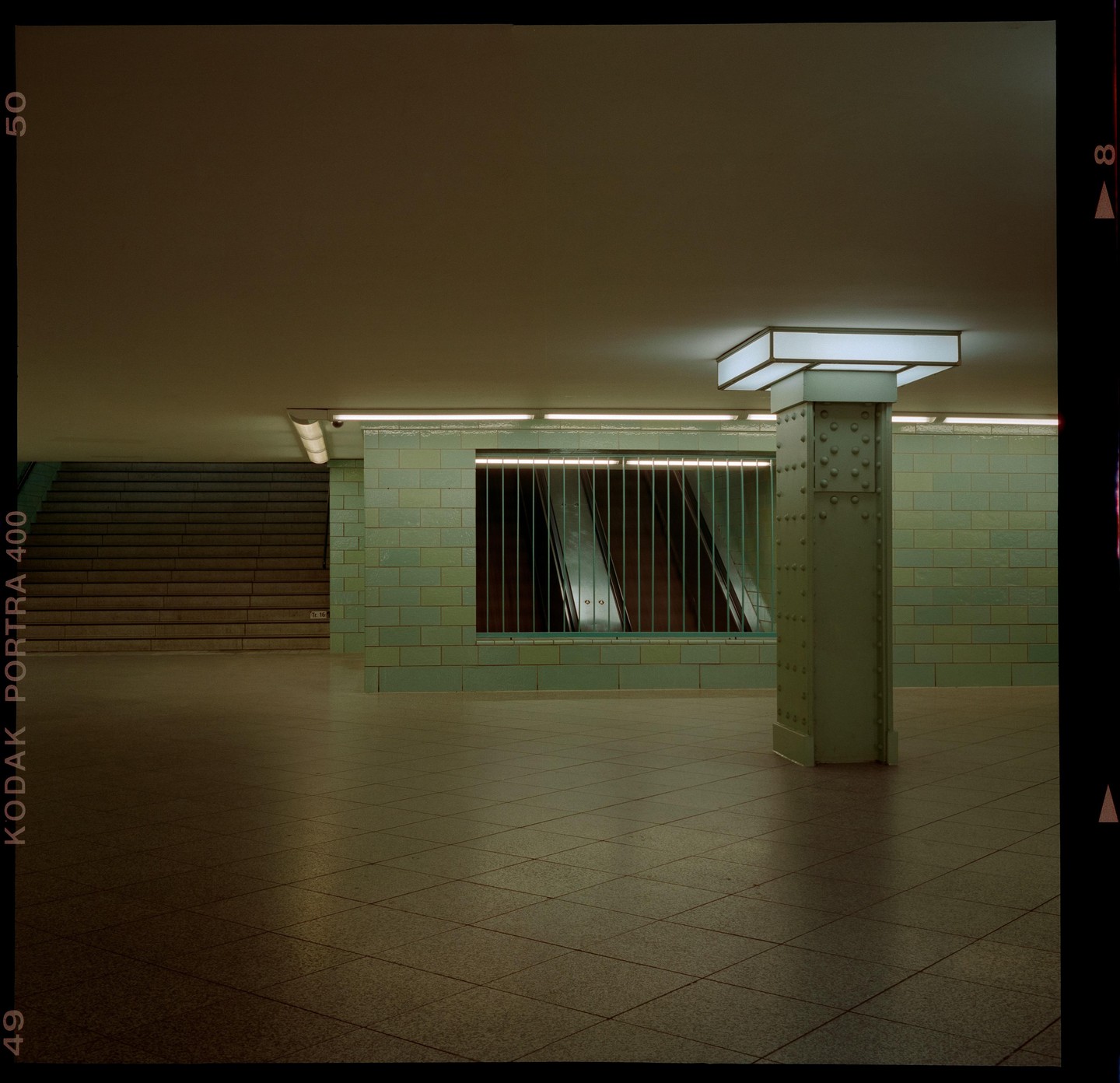A subway station features a tiled wall, a large pillar, and a set of stairs leading up.