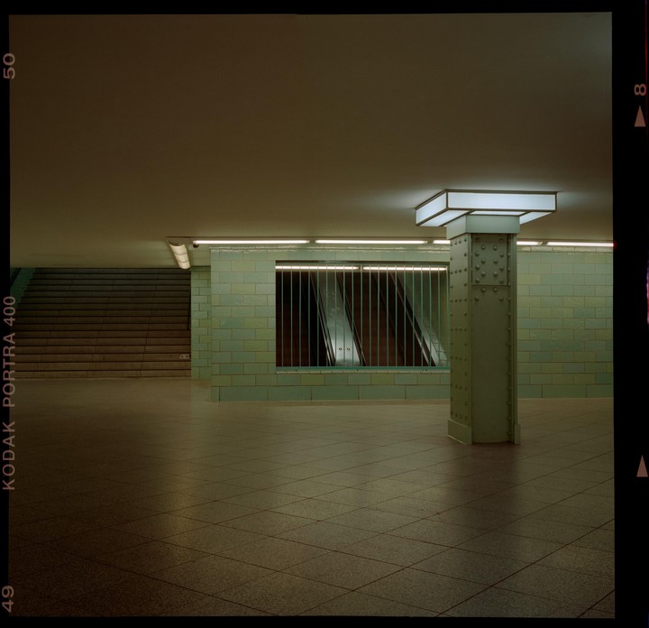 A subway station features a tiled wall, a large pillar, and a set of stairs leading up.