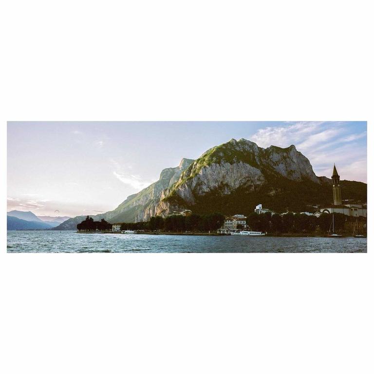 A panoramic landscape shows a body of water with mountains and buildings along the shore.