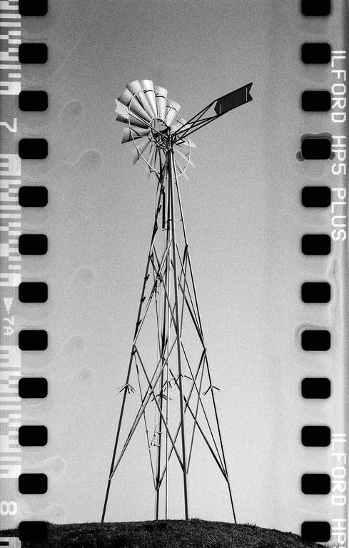 A black and white photograph shows a windmill on a grassy hill against a bright sky, with film borders visible on both sides.