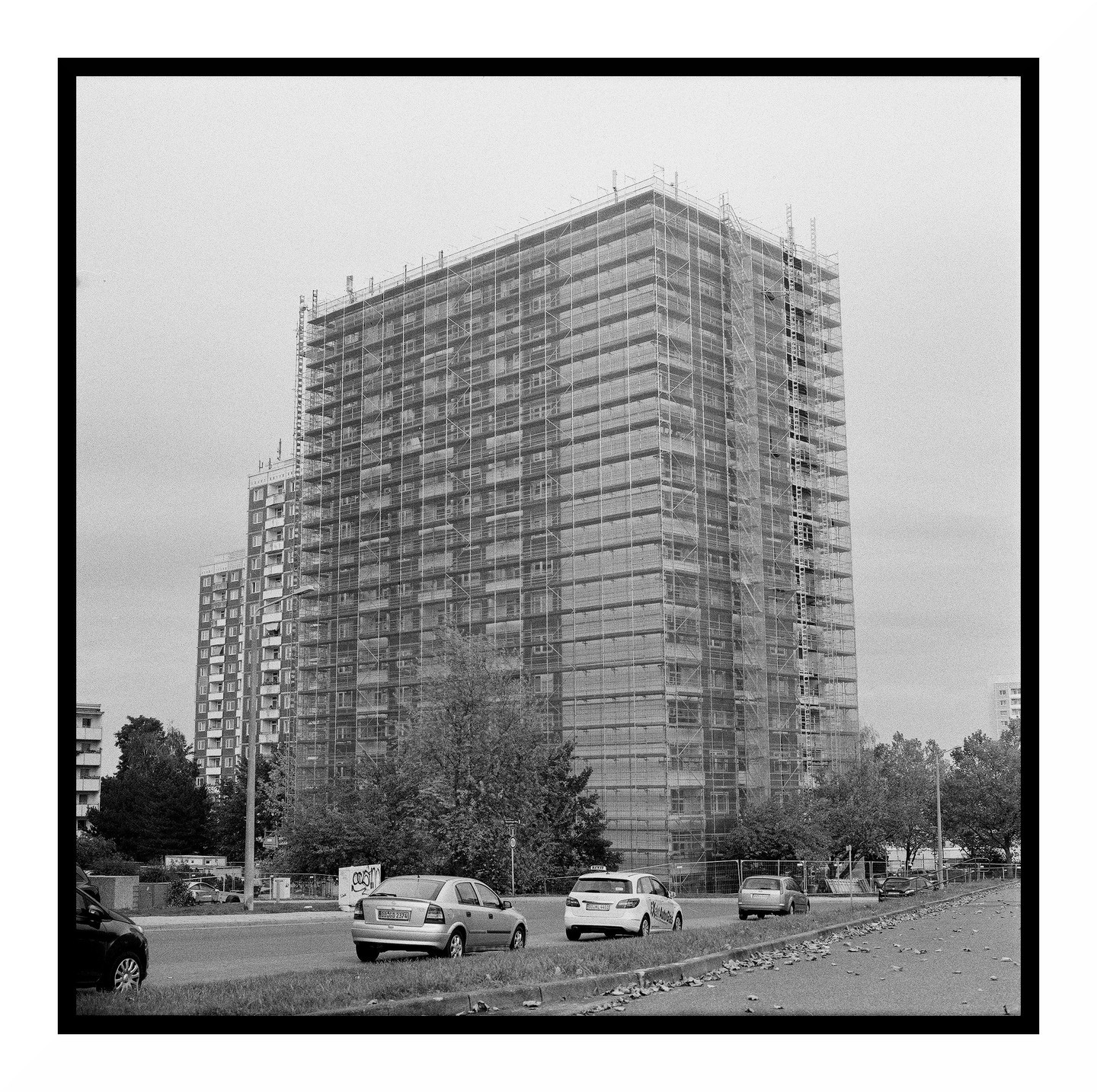 A tall building covered in scaffolding stands next to a street with cars and trees.
