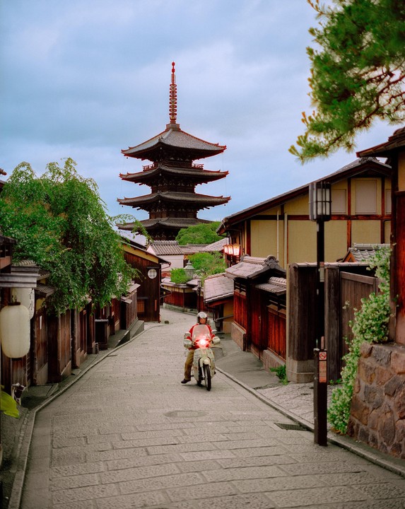 A person on a scooter drives up a cobblestone street lined with traditional buildings and a pagoda in the background.