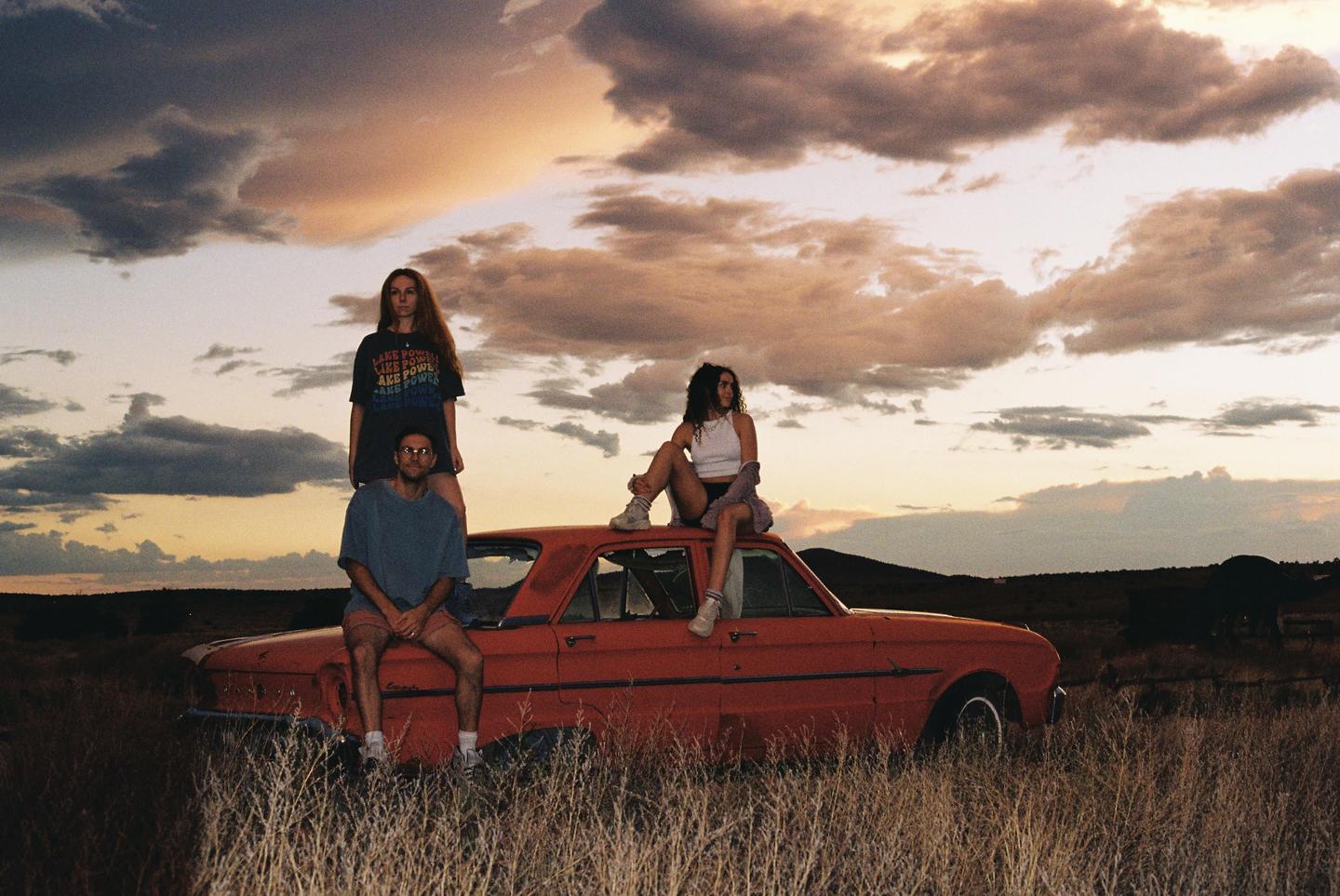 Three young people pose on and around an old red car in a field at sunset.