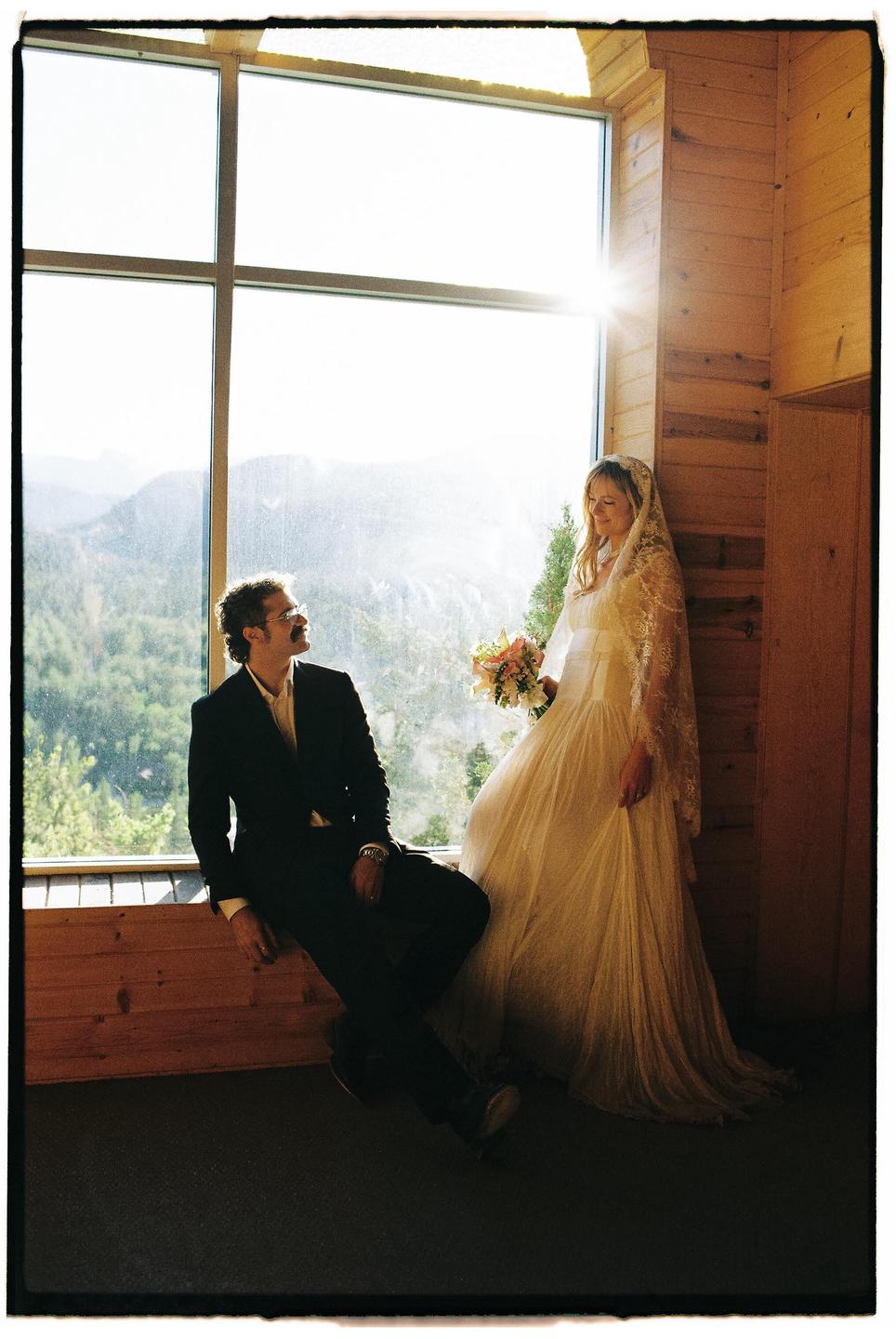 A couple poses for a wedding portrait in front of a large window overlooking a mountain landscape.