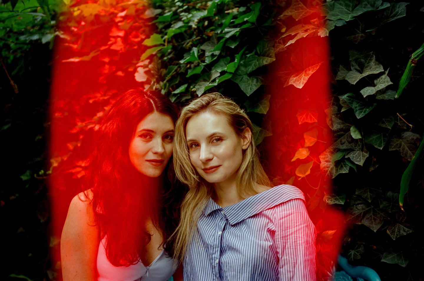 Two women pose for a portrait in front of an ivy-covered wall with a red light leak across the image.