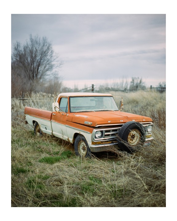 An old orange and white pickup truck is parked in a field of dry grass under a cloudy sky.