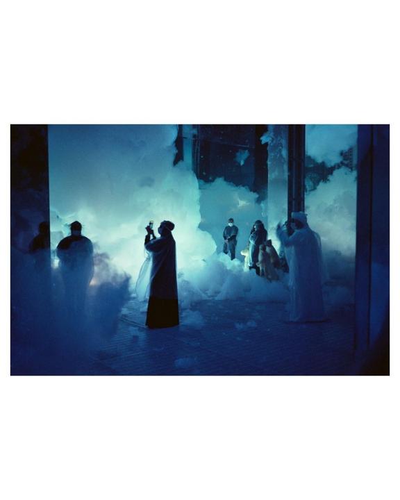 Several people stand among blue-lit clouds of mist in a dark room.