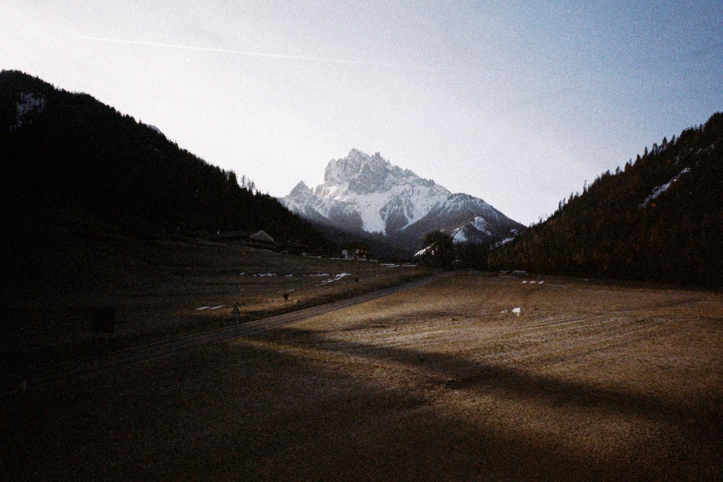 A snow-capped mountain peak rises above a valley with a road and fields, flanked by dark, forested hills.