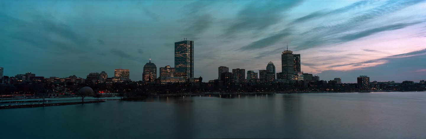 A panoramic view of the Boston skyline at dusk, with city lights reflecting on the water.