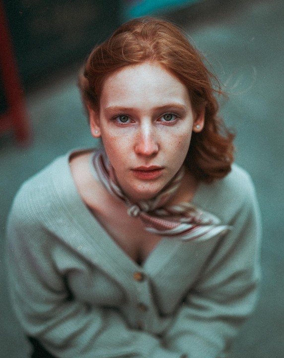 A woman with red hair and freckles looks up at the camera, wearing a light-colored cardigan and a patterned scarf.