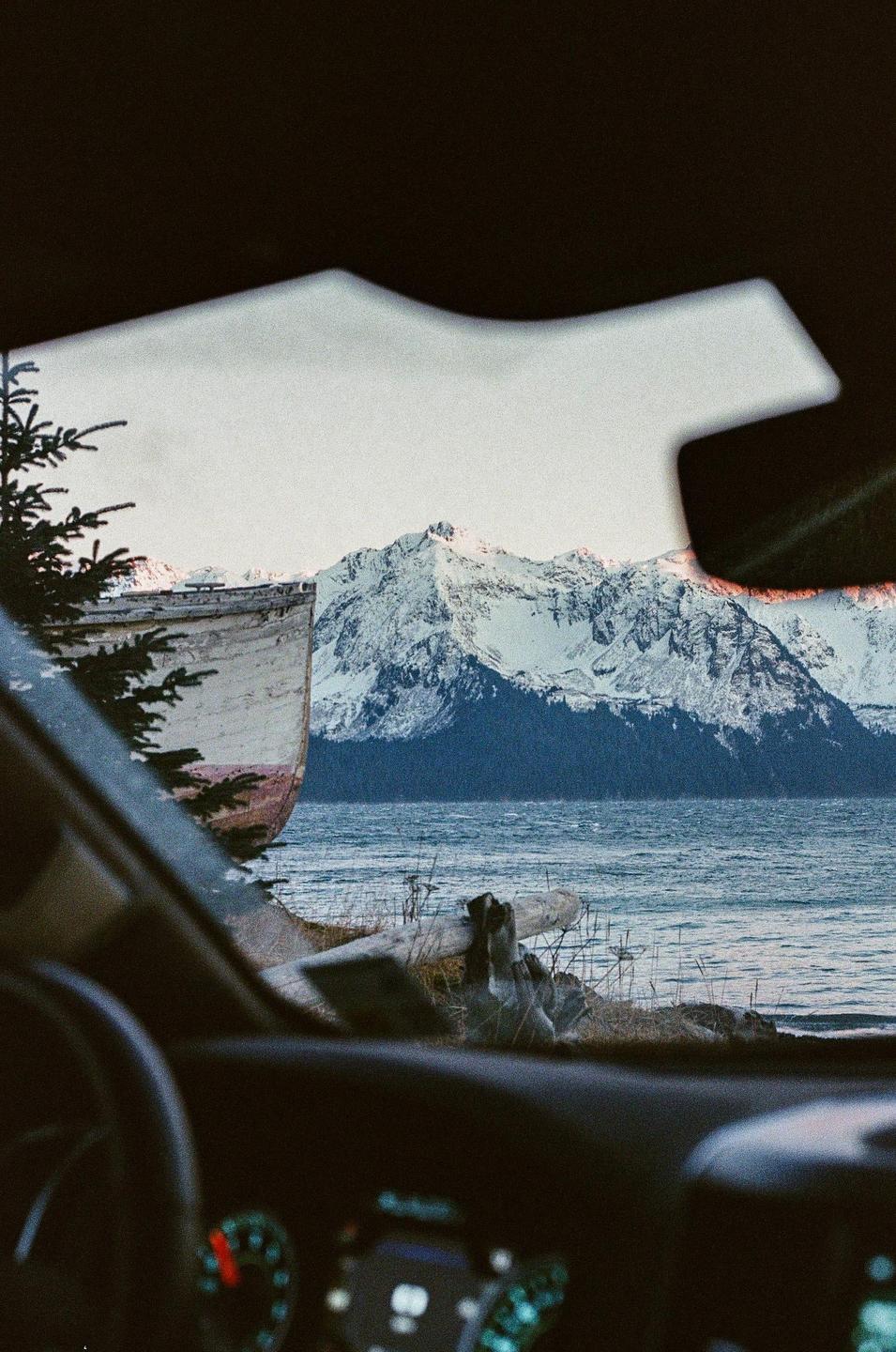 A view from inside a car shows a boat on the shore of a lake with snow-capped mountains in the background.