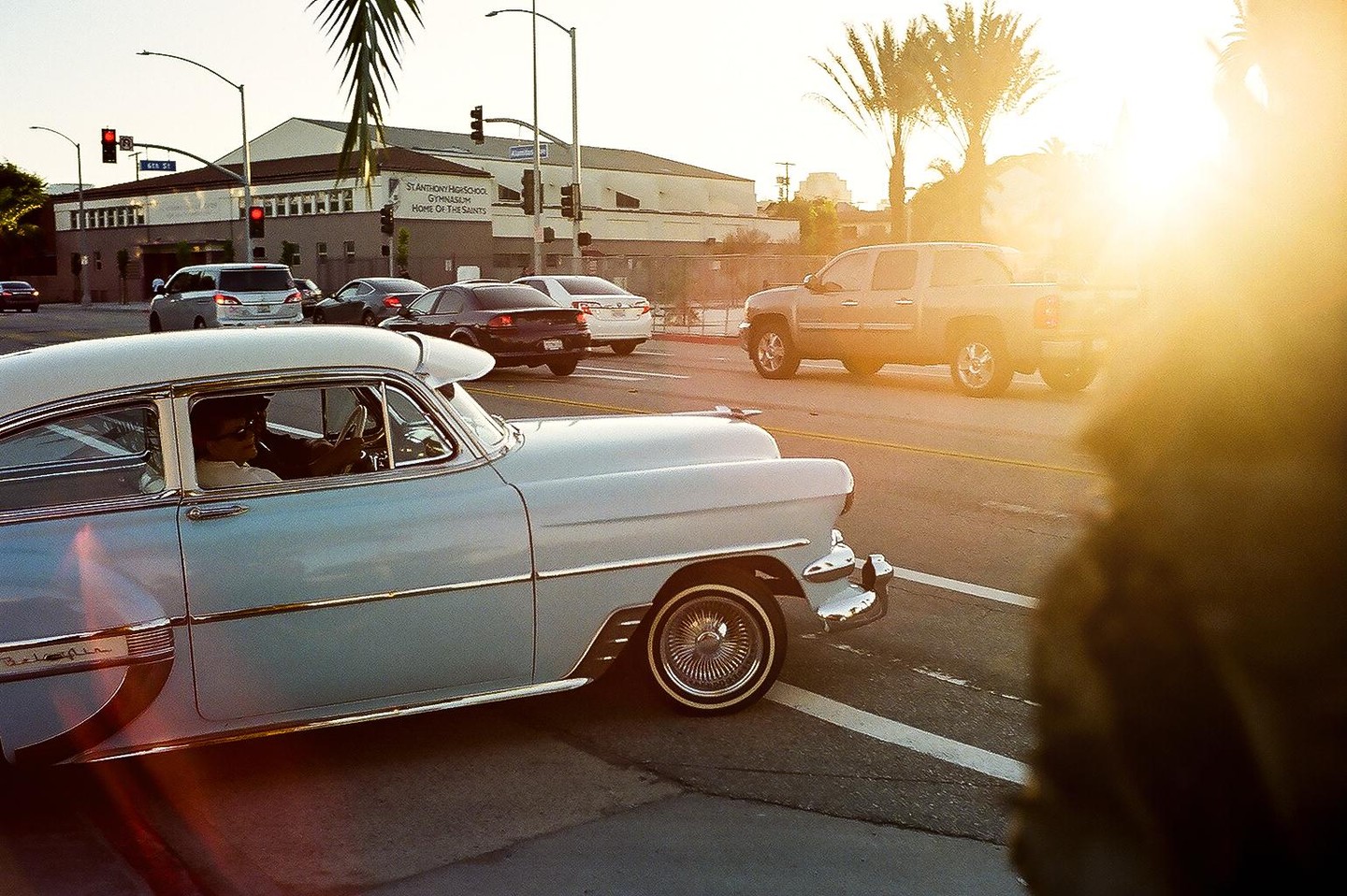 A light blue vintage car with a man driving is on a street with other vehicles and a building in the background.