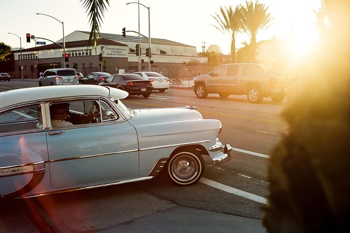 A light blue vintage car with a man driving is on a street with other vehicles and a building in the background.