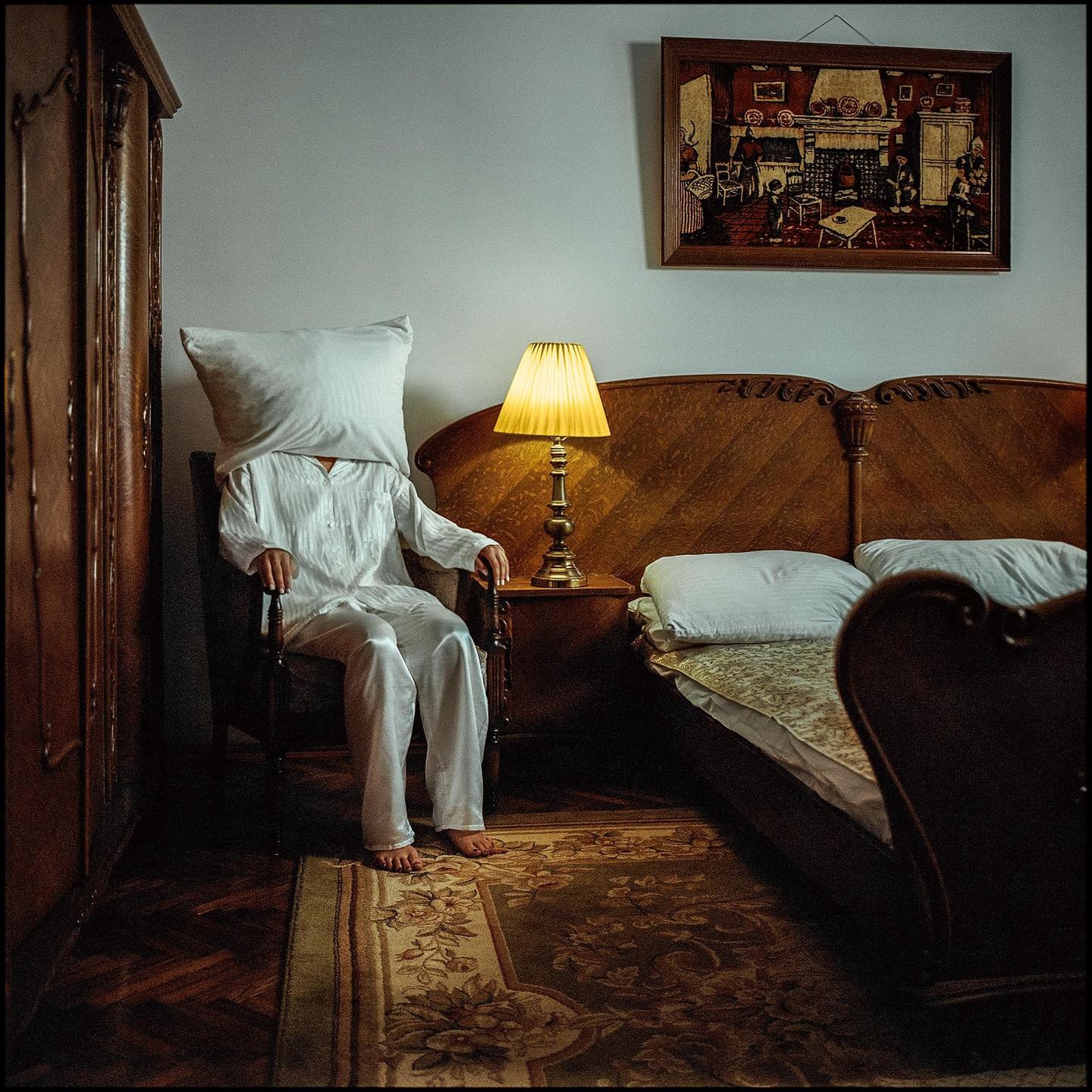 A person wearing white pajamas sits in a chair with a pillow over their head next to a bed and a lit lamp.