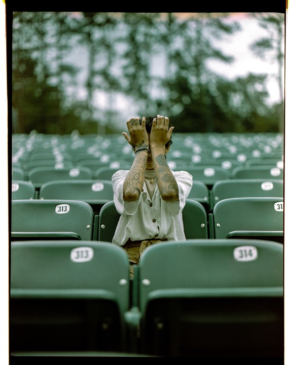 A person with tattooed arms covers their face with their hands while sitting in a stadium seat.