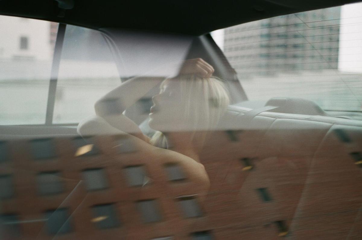 A woman with blonde hair is visible through a car window, with reflections of buildings superimposed on the image.