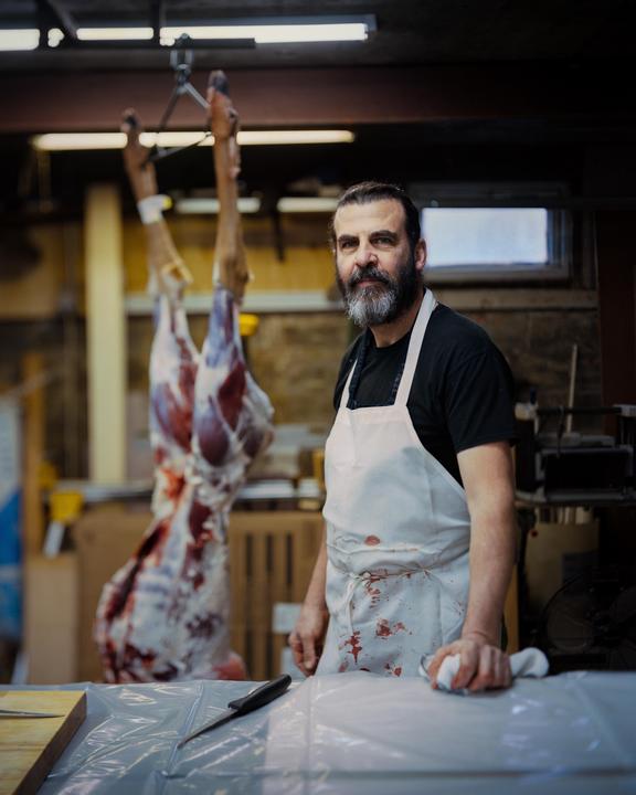 A man wearing a blood-stained apron stands in a garage with a hanging animal carcass in the background.