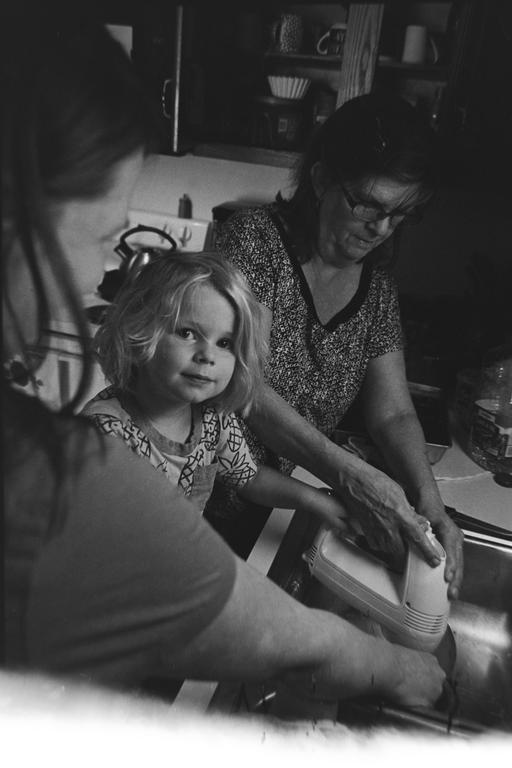 A child looks at the viewer while a woman washes a hand mixer in a kitchen sink.
