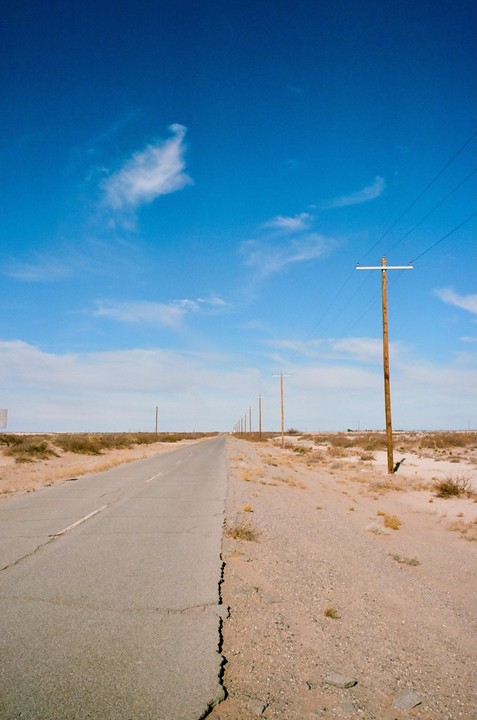 A long, cracked road stretches into the distance, lined by telephone poles and dry desert brush under a bright blue sky.