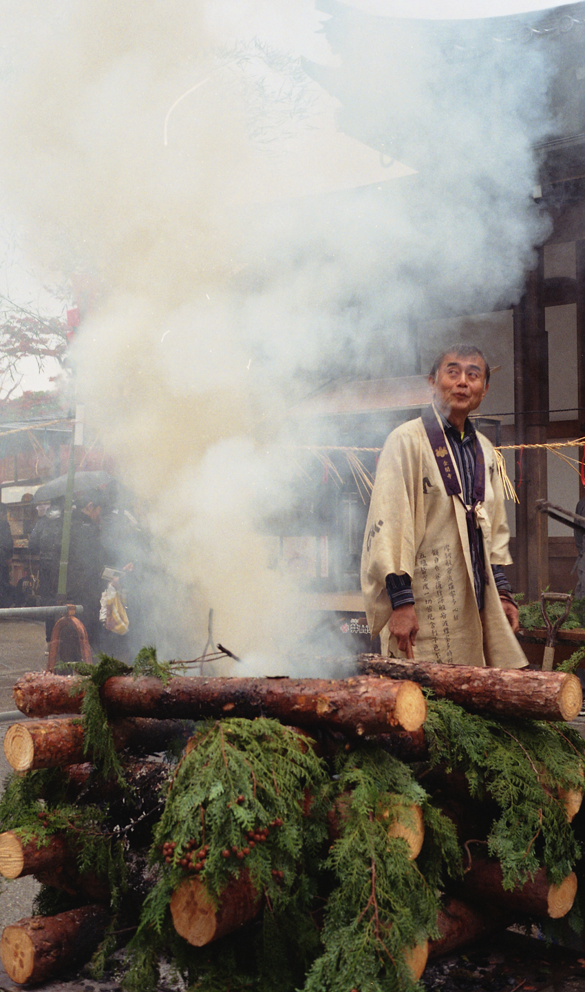 A man in traditional clothing stands next to a smoking pile of logs and greenery.