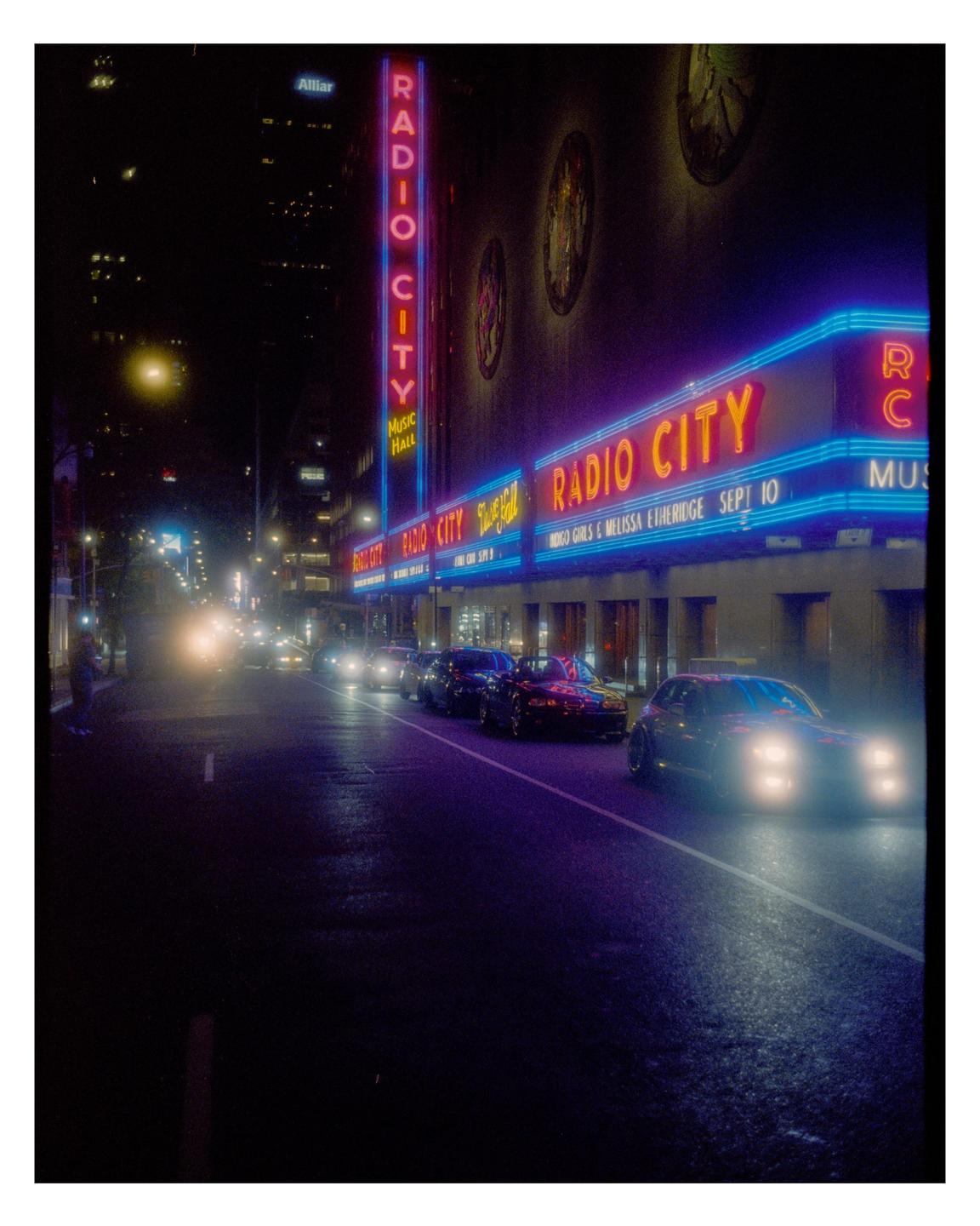 A street at night is illuminated by the neon signs of Radio City Music Hall and the headlights of cars.