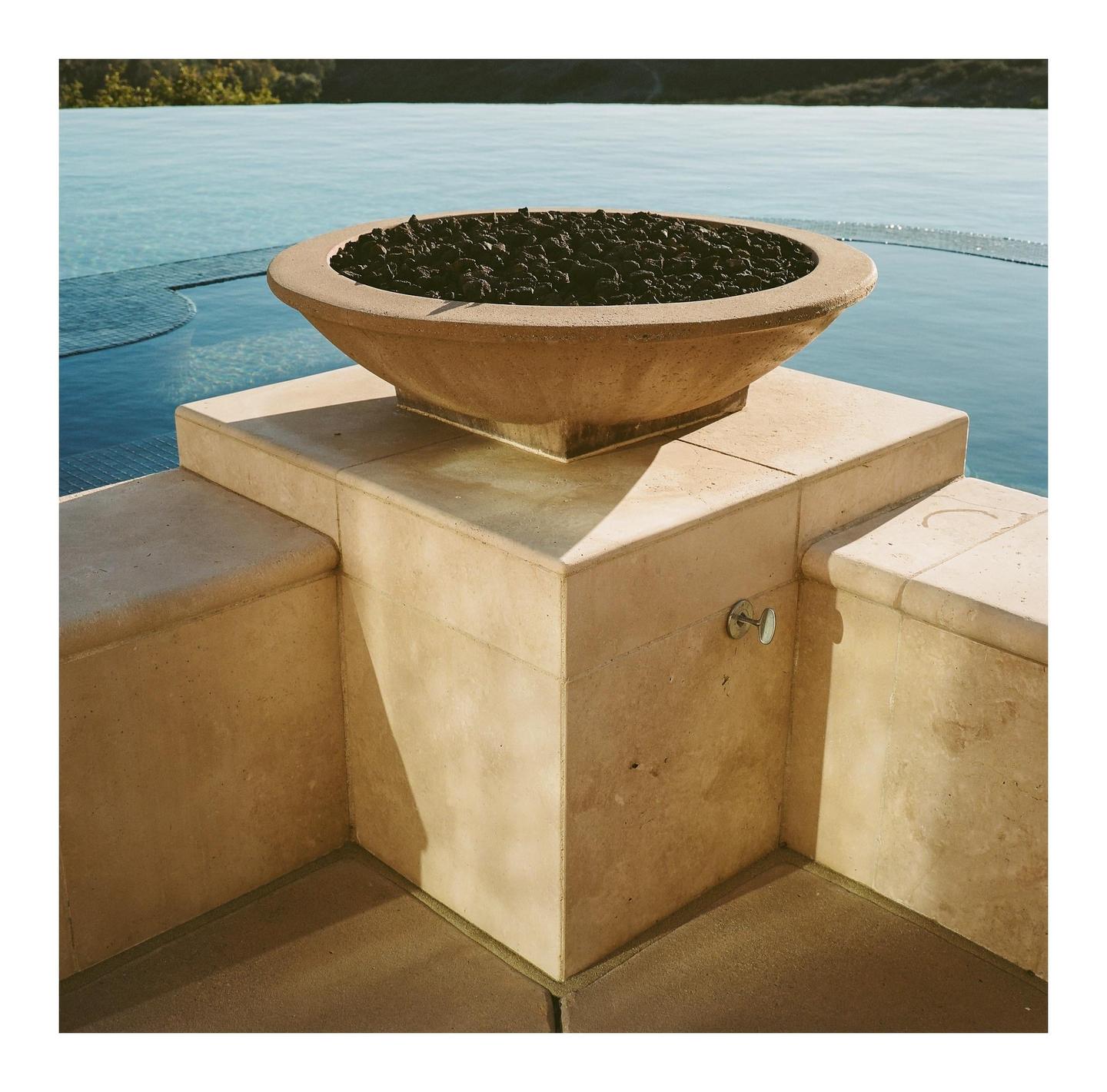 A concrete fire pit filled with dark rocks sits on a stone ledge next to a swimming pool.
